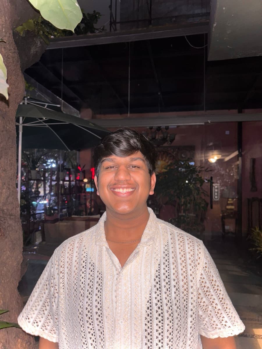 Kartikey smiling, in a white crochet shirt, at night outside a café